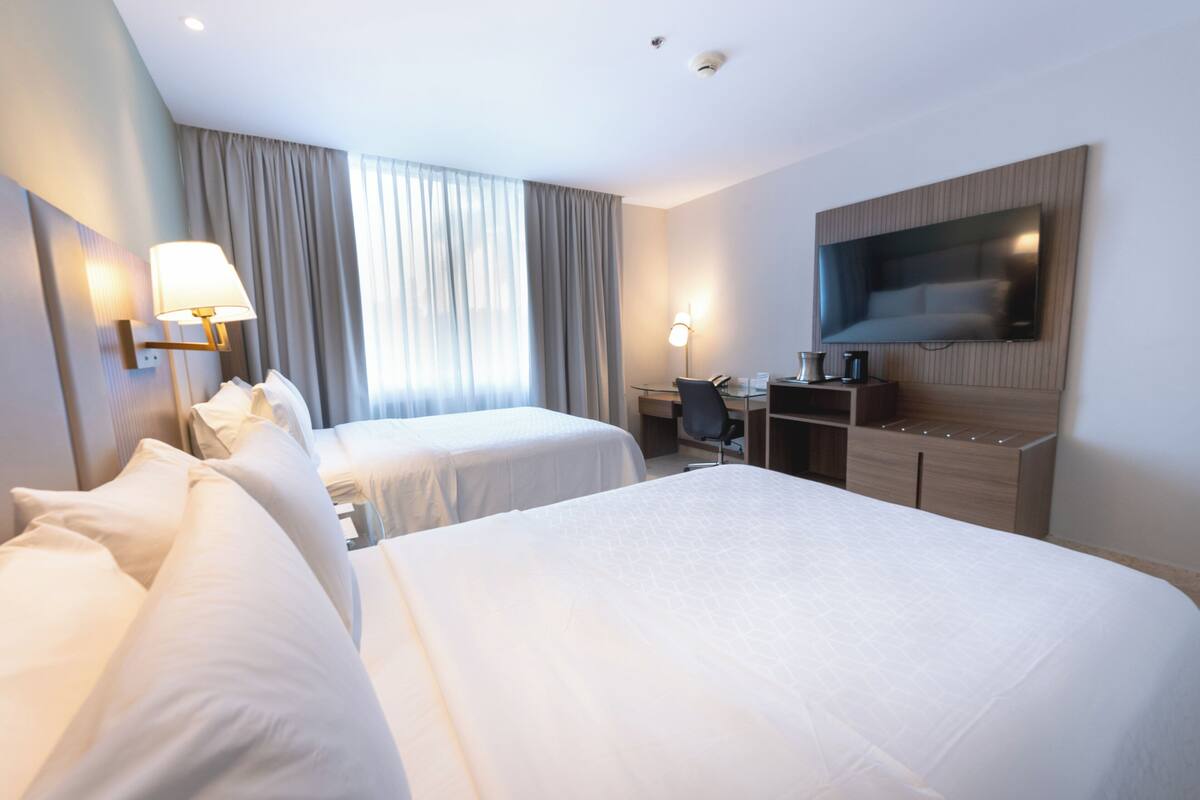 A hotel room with two beds, a desk, a chair, a flat-screen TV, and a large window with curtains.