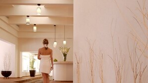 Un ambiente de spa con un camino de piedra, un banco de madera y una persona con bata blanca caminando hacia una recepción adornada con un jarrón de flores.