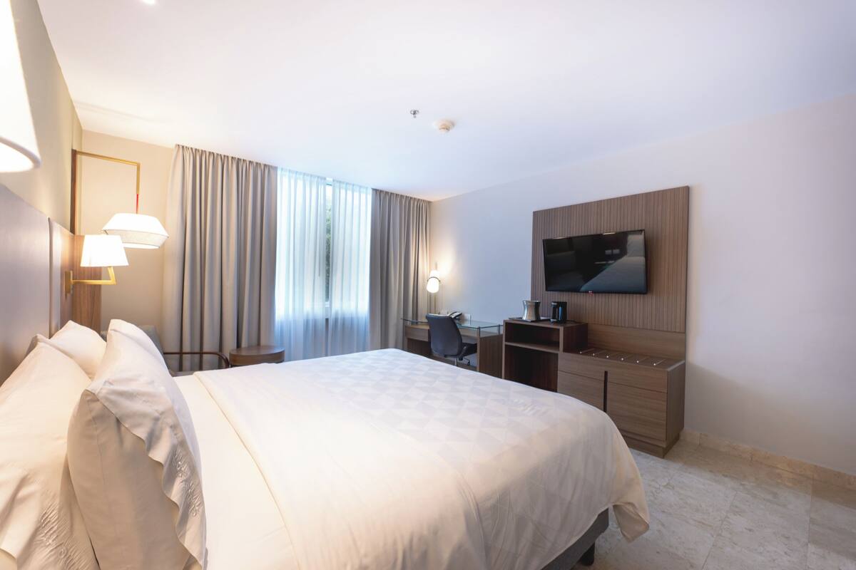 A hotel room with a large bed, a desk with a chair, a television mounted on the wall, and a window with curtains.