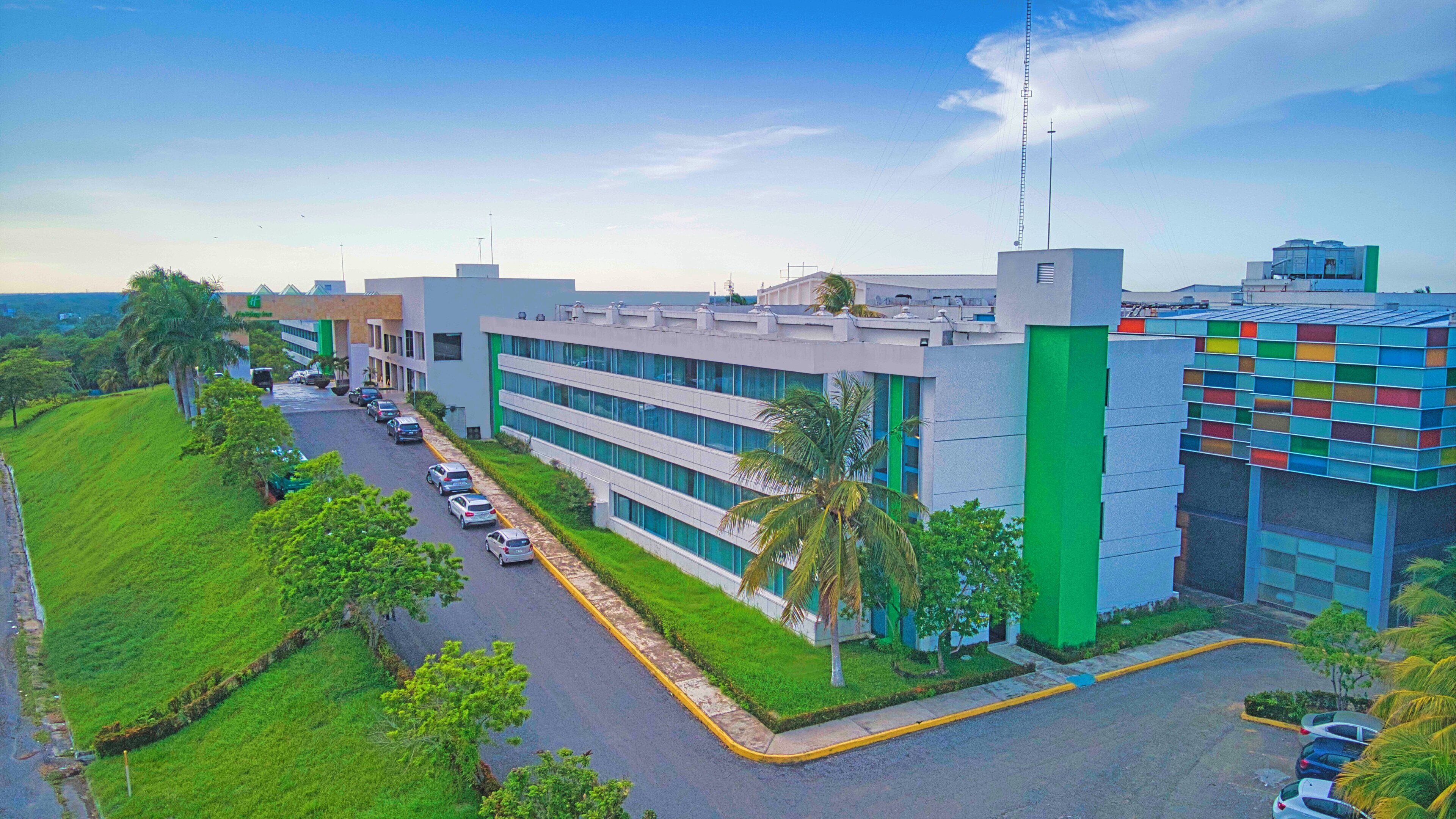 Un edificio moderno con un largo camino de entrada bordeado de coches aparcados, rodeado de vegetación y palmeras.