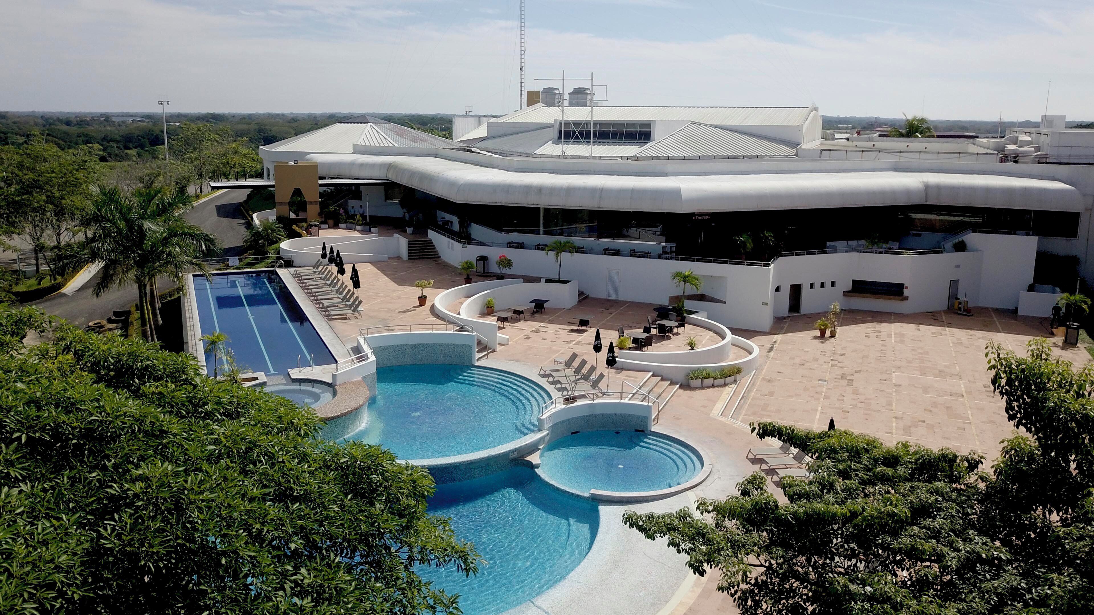 A modern hotel with a rooftop pool area, including a large swimming pool and a smaller circular pool, surrounded by lounge chairs and potted plants.