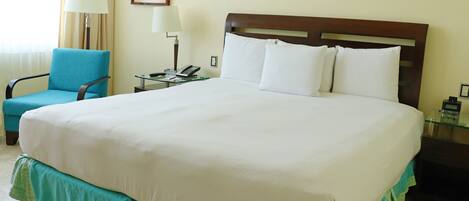 Una habitación de hotel con una cama grande, un sillón azul, una mesita de noche con una lámpara y un cuadro enmarcado en la pared.