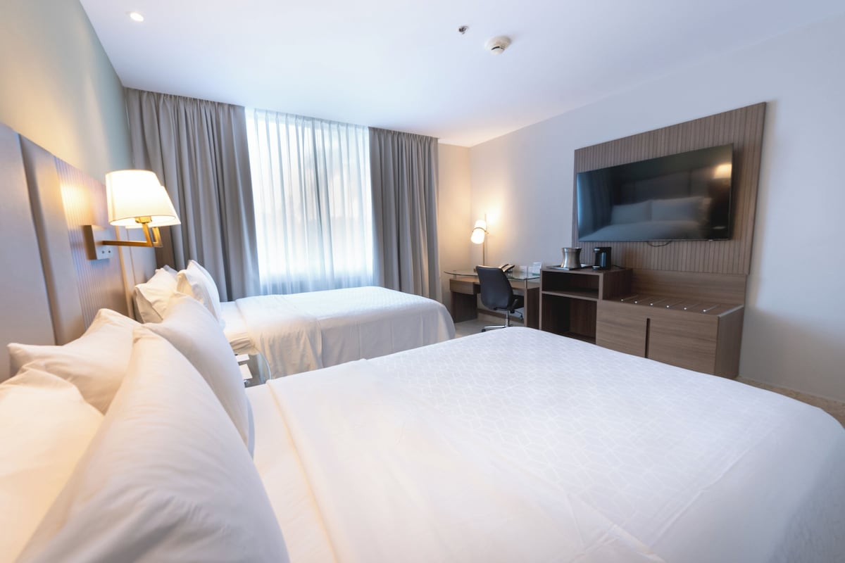 Habitación de hotel con dos camas, un escritorio, una silla, un televisor de pantalla plana y un ventanal grande con cortinas.