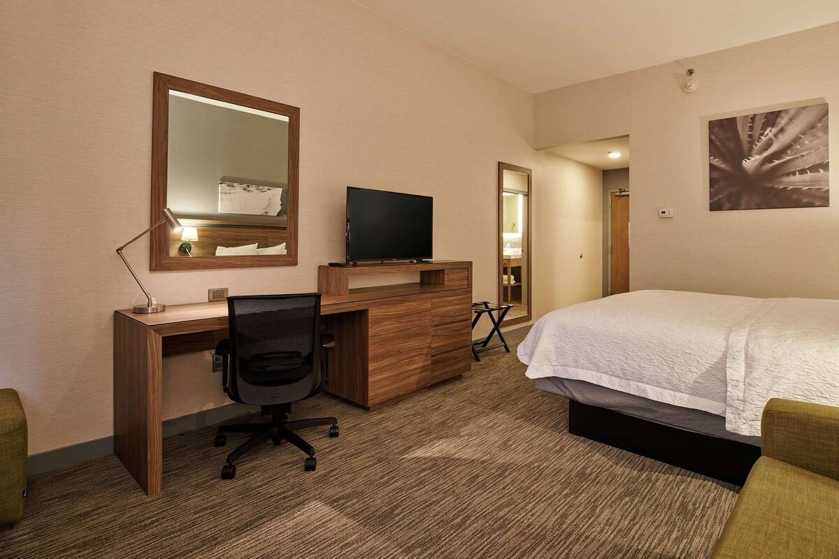 A hotel room with a bed, a desk with a chair, a TV, a mirror, and a bathroom visible through a door.