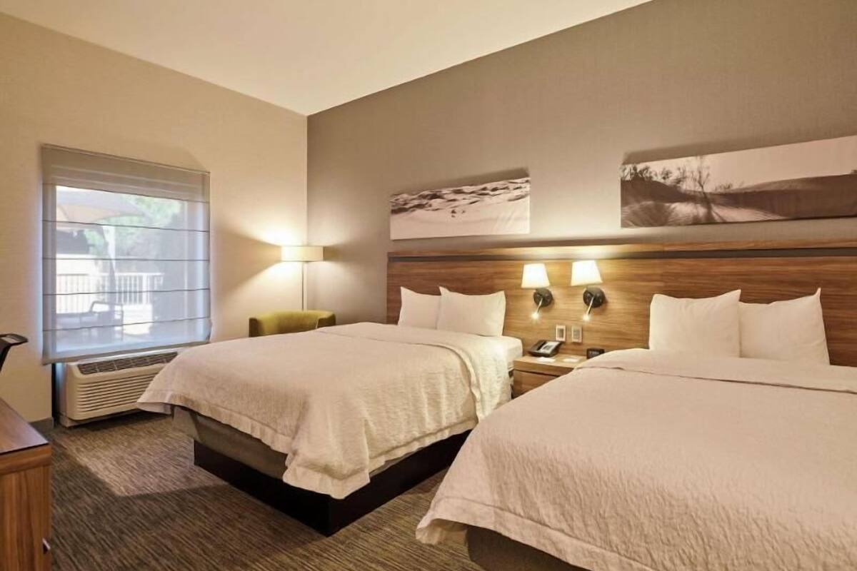 A hotel room with two beds, a window with blinds, a wall-mounted lamp, and a painting.