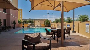 Un espace extérieur avec une piscine, des chaises longues et des parasols, entouré de bâtiments et surplombé par un ciel dégagé.