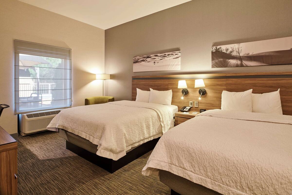 A hotel room with two beds, a window with blinds, a lamp, and a wall-mounted artwork.