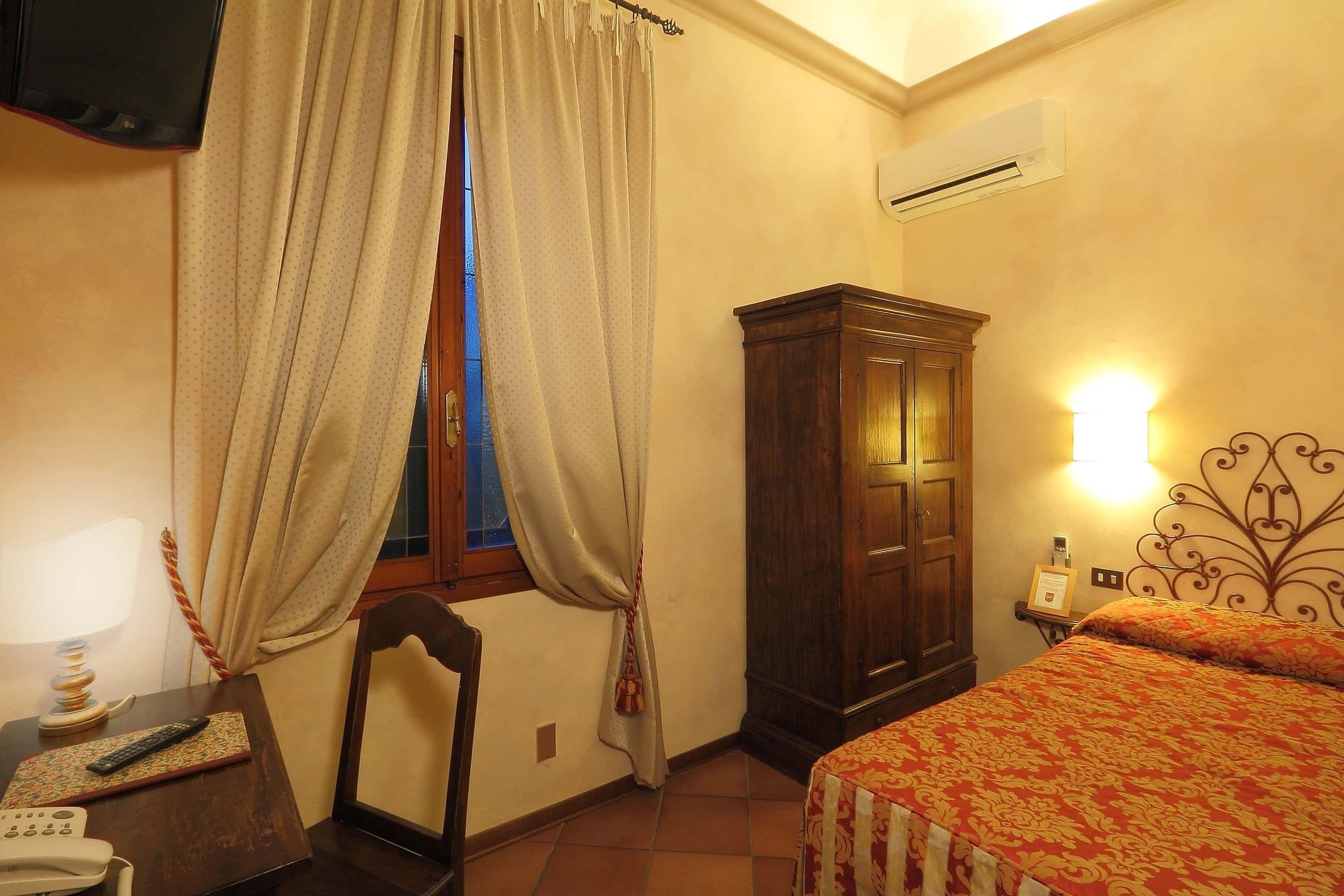 Une chambre à coucher avec un lit, une armoire en bois, un bureau avec une lampe, un téléphone et une fenêtre avec des rideaux.
