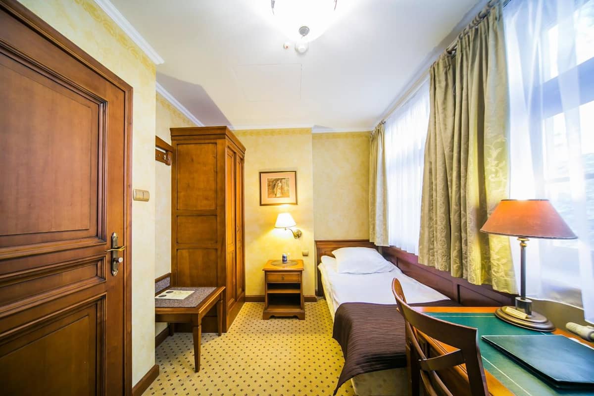 A hotel room with a wooden wardrobe, a bedside table with a lamp, a bed with a brown headboard, a chair, and a window with curtains.