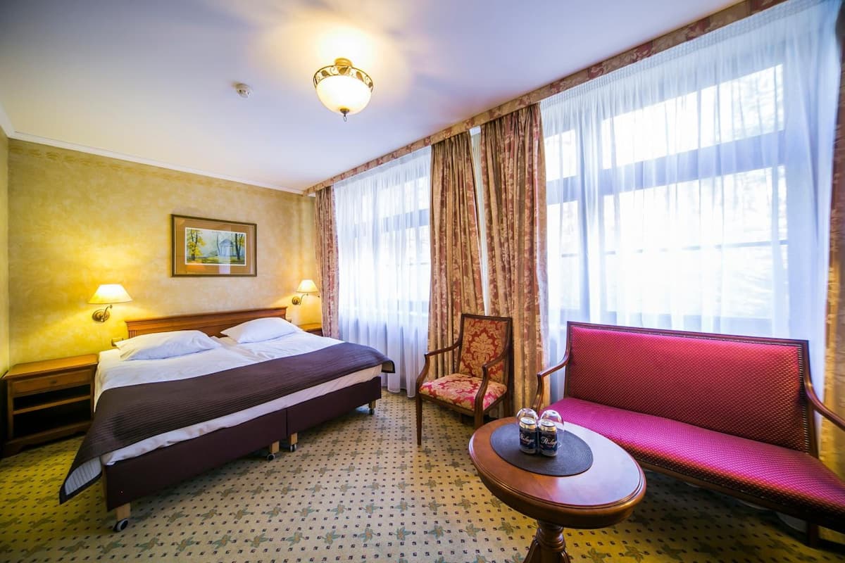 A hotel room with a double bed, two bedside tables with lamps, a red sofa, a small round table with two bottles, and a large window with sheer curtains.