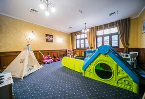 Zona de juegos con un tipi, un tobogán y un túnel. Se ve una ventana con cortinas y una araña.