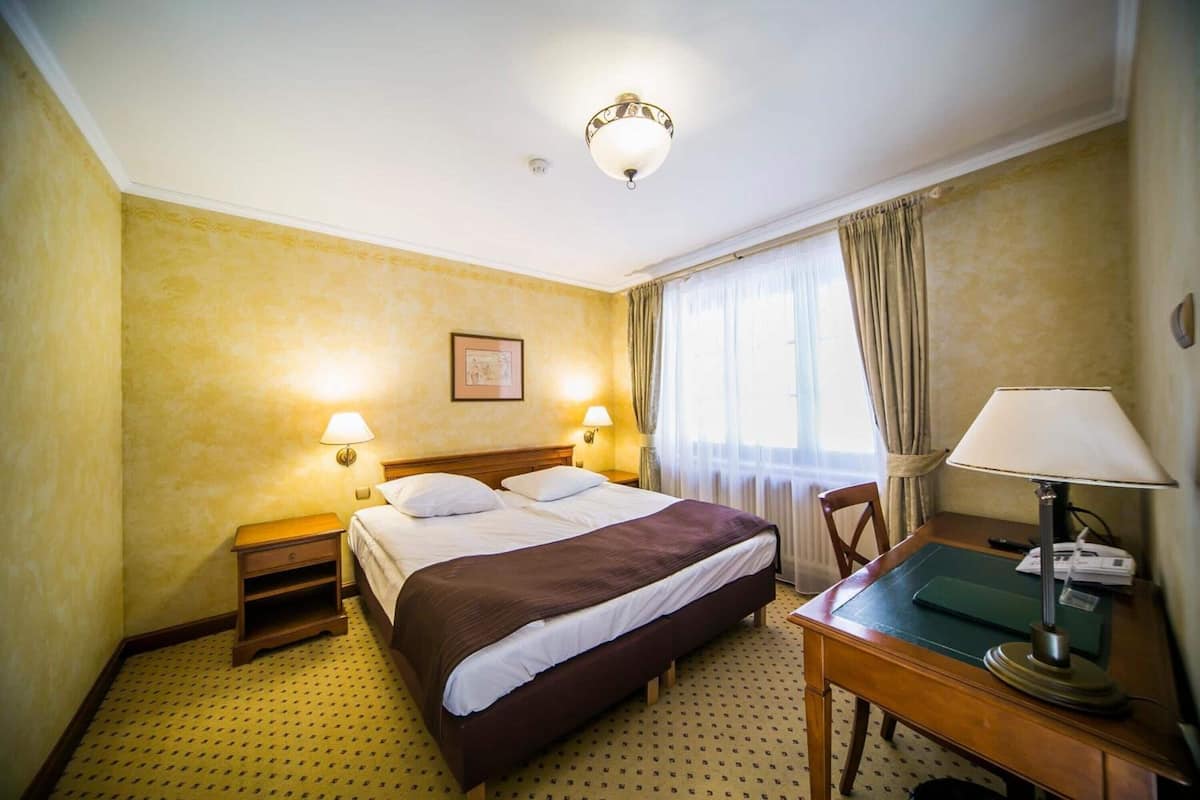 A hotel room with a bed, bedside lamps, a desk with a lamp, and a window with curtains.