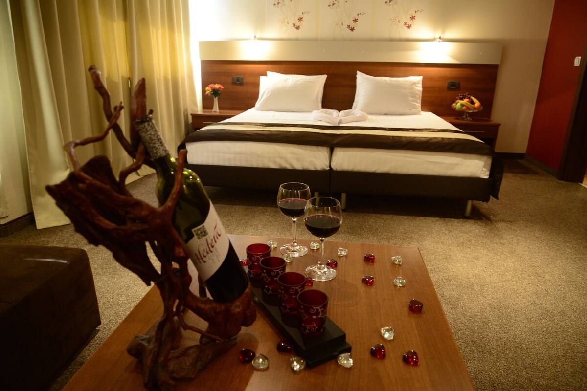 Habitación de hotel con cama, mesitas de noche, una mesa de centro de madera con copas de vino y una botella, y elementos decorativos.