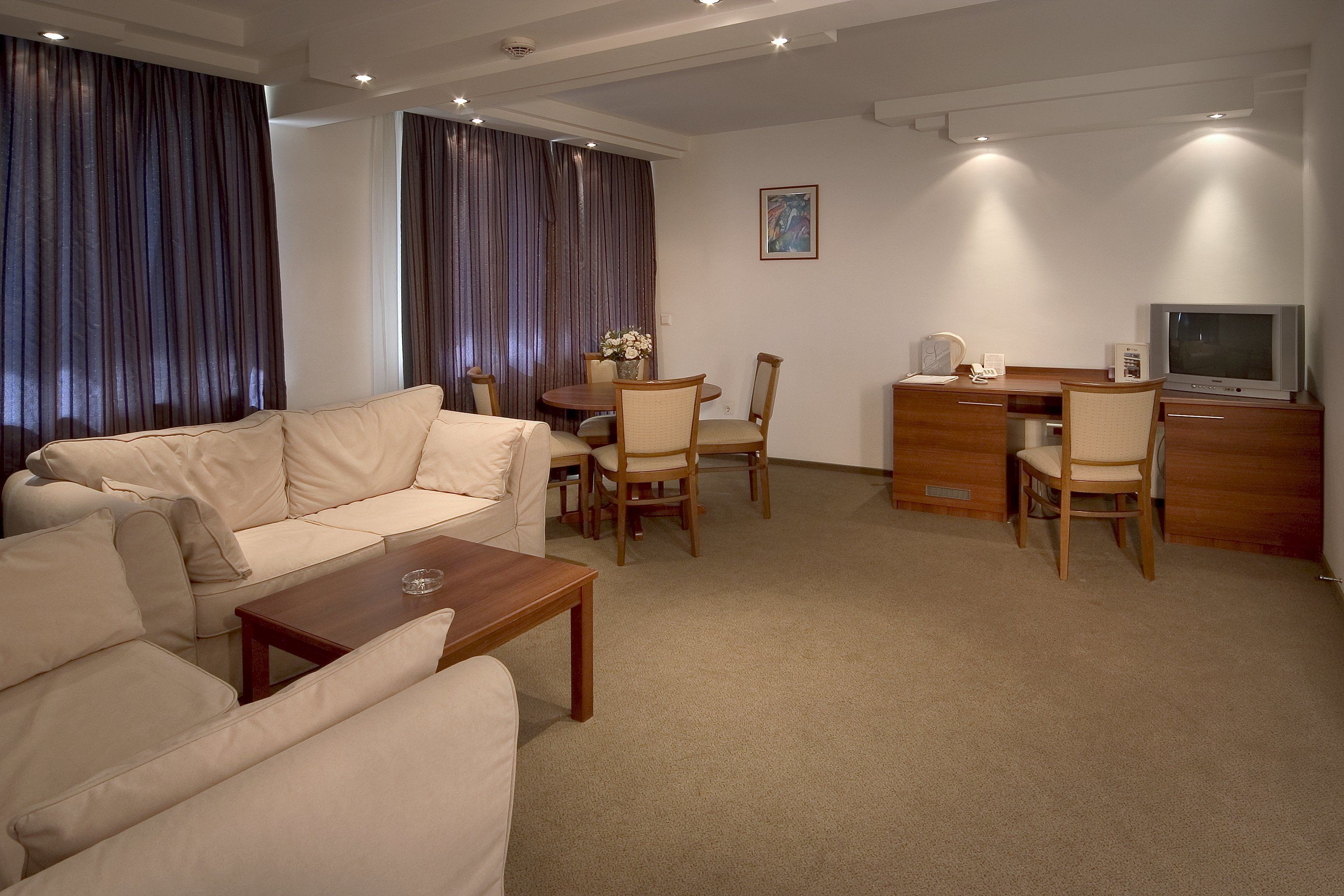 Una habitación de hotel moderna con un sofá color beige, un escritorio de madera, un televisor y una mesa de comedor con sillas.