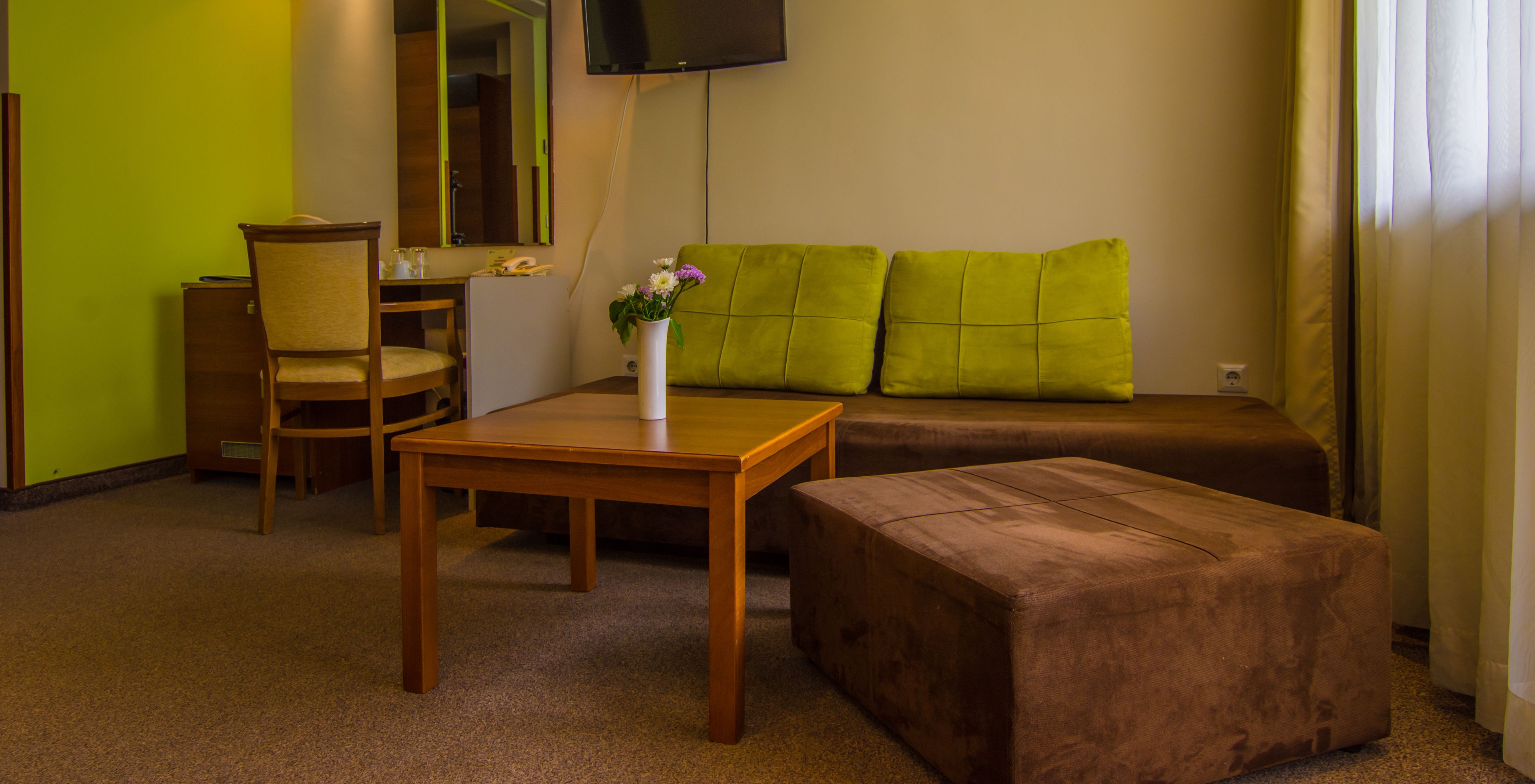 A hotel room with a wooden desk, a green sofa, a brown ottoman, a chair, and a small table with a vase of flowers.