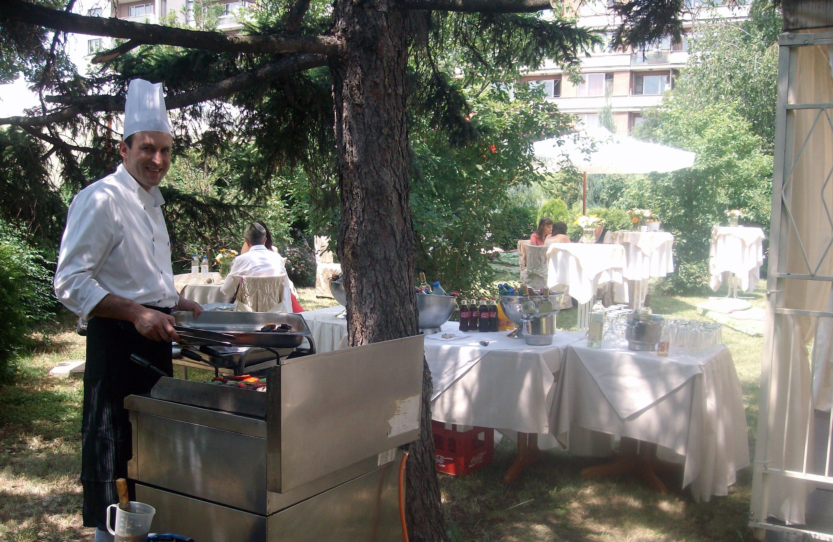 Ein Koch bereitet im Freien Essen zu, mit einem Grill und Kochgeschirr. Im Hintergrund sind Tische mit weißen Tischdecken und verschiedene Speisen zu sehen.