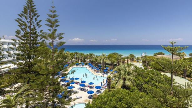 A resort pool area with multiple sunbathing spots and umbrellas, surrounded by palm trees and overlooking a beach.