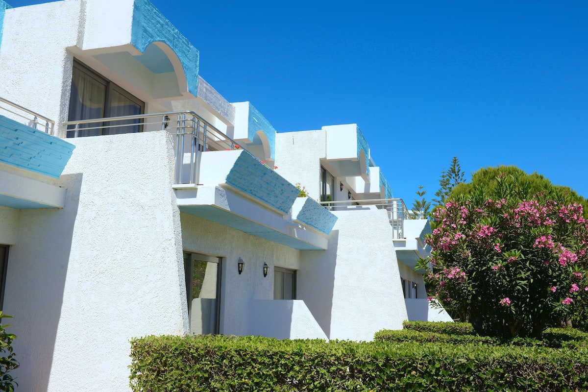Un edificio moderno con paredes exteriores blancas y detalles azules, con balcones y un jardín bien cuidado con flores rosadas.