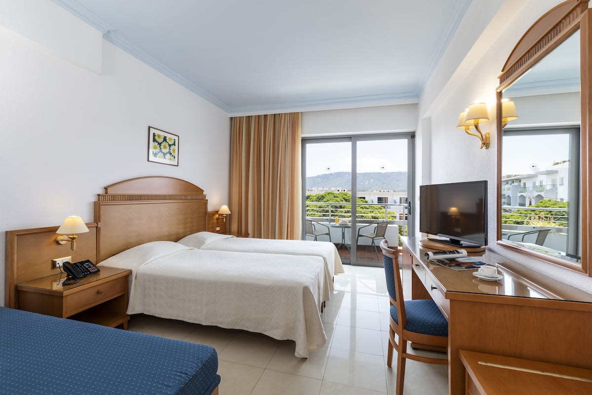 Habitación de hotel con dos camas, un escritorio con televisor, teléfono y balcón con vistas.