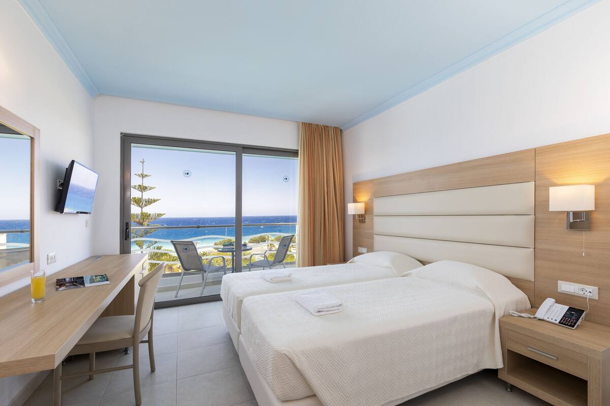 Habitación de hotel con dos camas, un escritorio con silla, televisor y vista al océano.