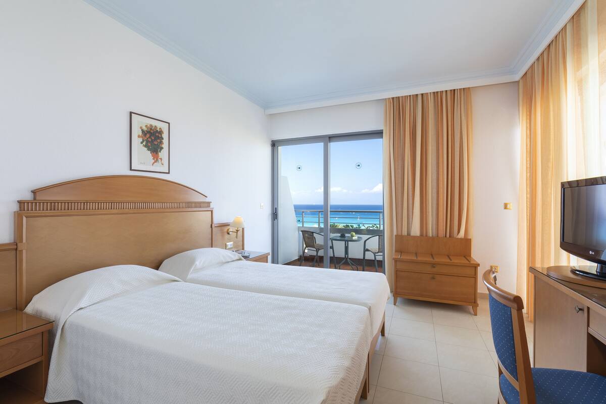 Habitación de hotel con dos camas, televisión, escritorio, silla y balcón con vista al océano.