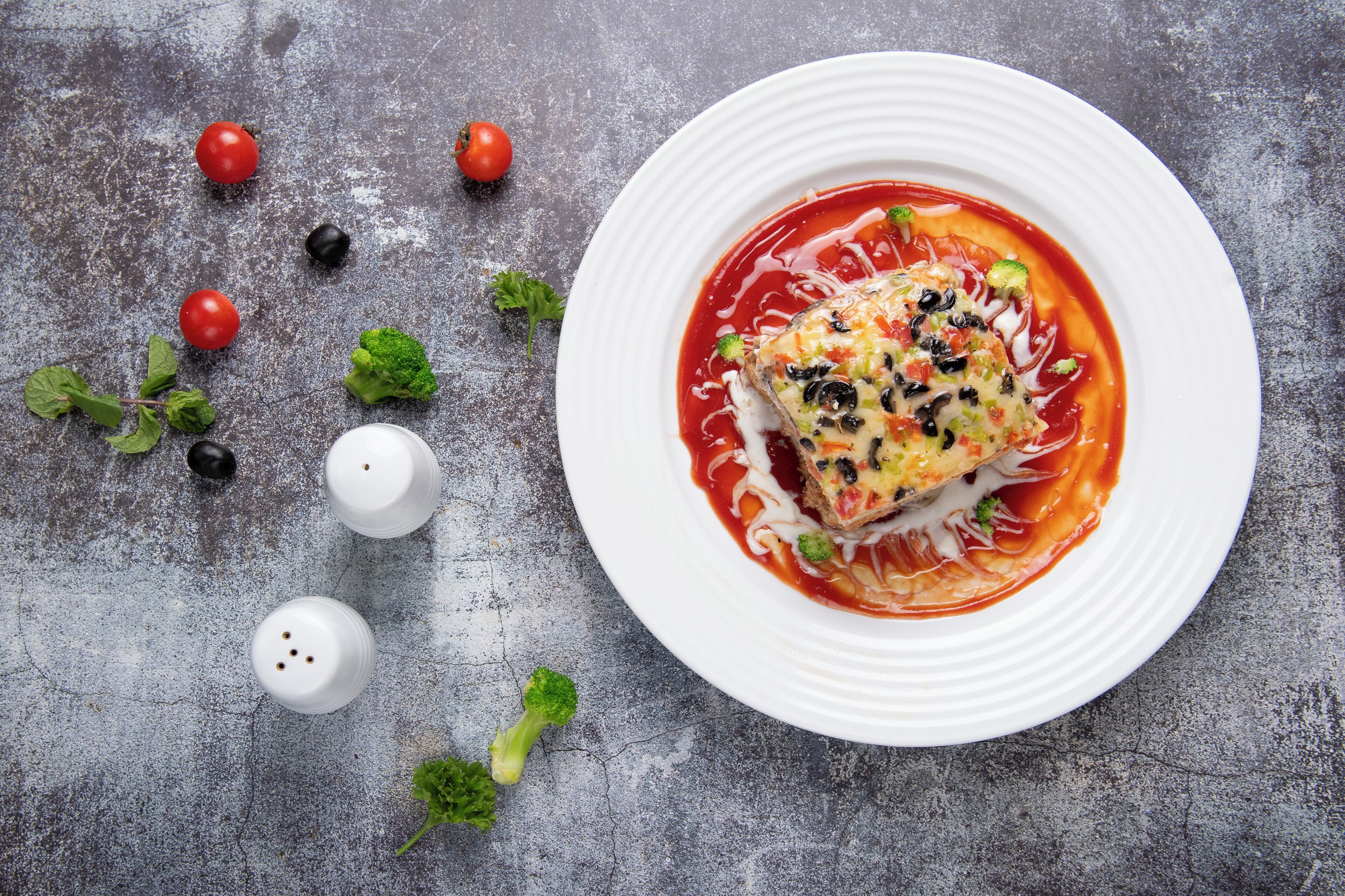 A white plate with a fish fillet on a bed of sauce, surrounded by scattered cherry tomatoes, broccoli, and black olives on a textured grey surface.