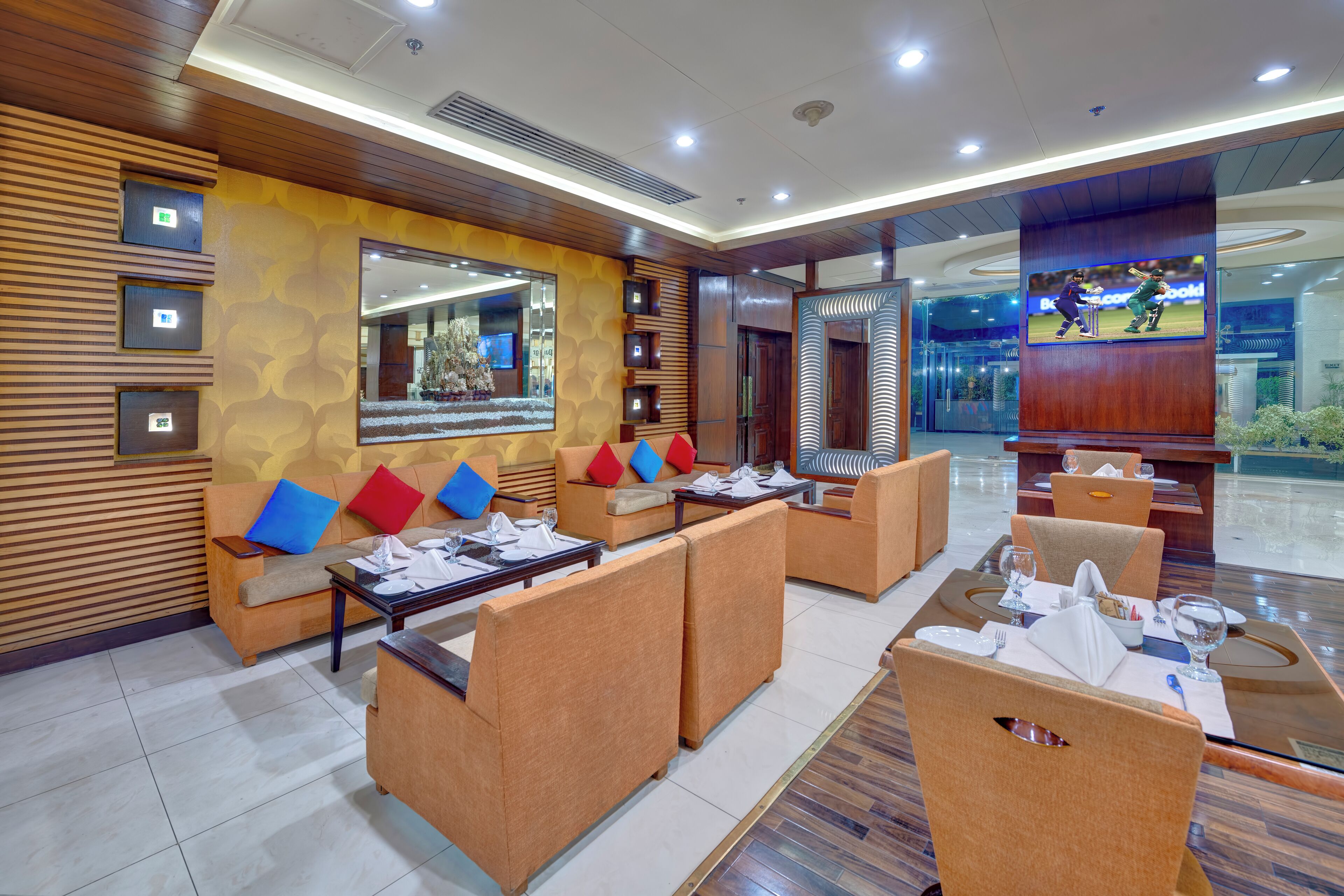 A modern hotel lobby with seating areas, a large mirror, and a television displaying a cricket match.
