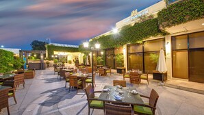 An outdoor dining area with wicker furniture and glass tables, surrounded by large windows and a building with a sign reading "Pearl Continental". - Pearl Continental Rawalpindi (Rawalpindi)
