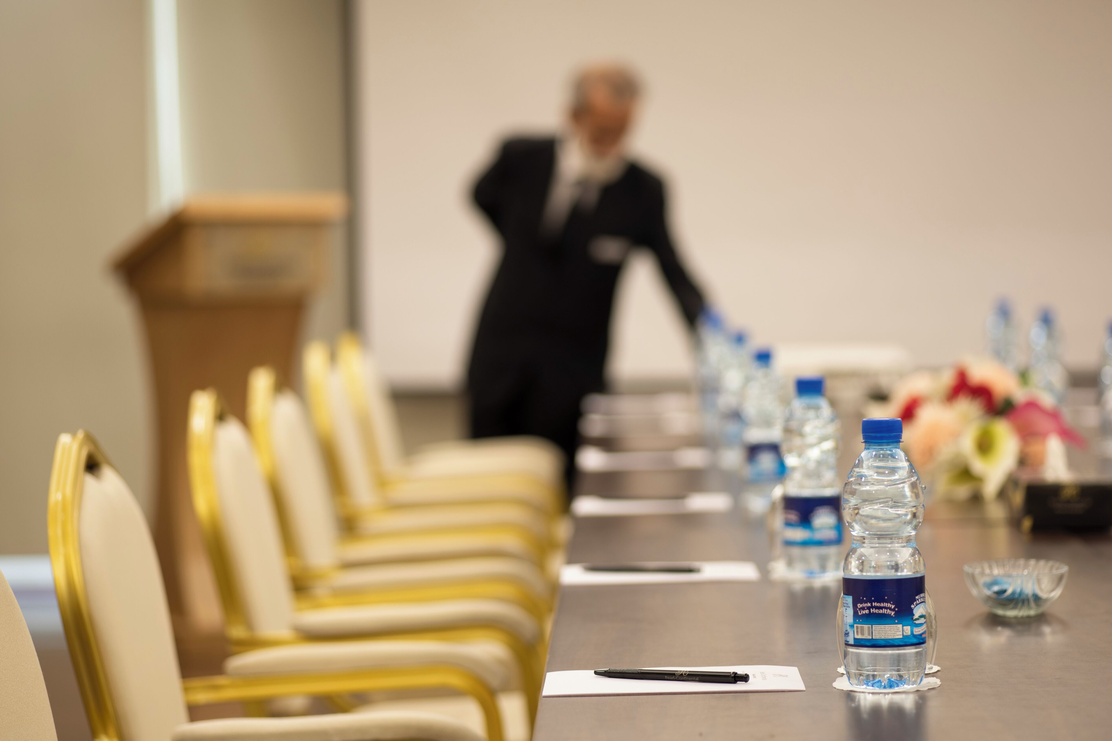 Een vergaderruimte met rijen gele stoelen, een spreekgestalte, waterflesjes en een pen op tafel.