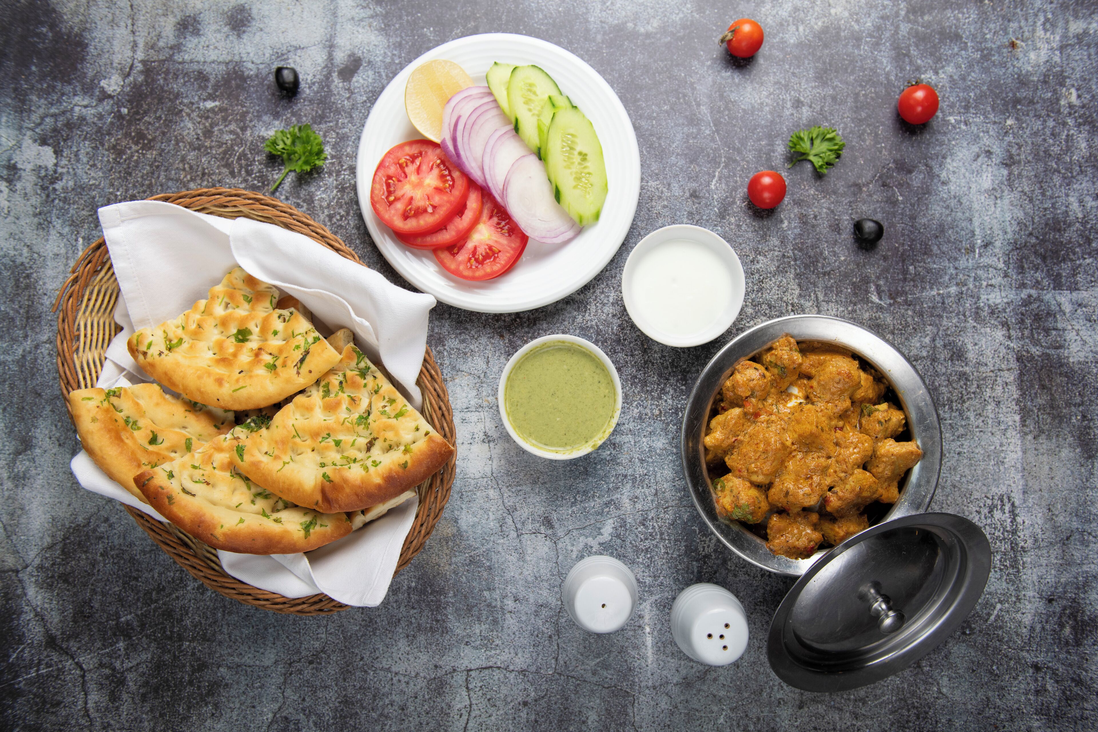 A table with naan bread, sliced tomatoes, cucumbers, onions, a bowl of curry, and two small bowls of sauce.