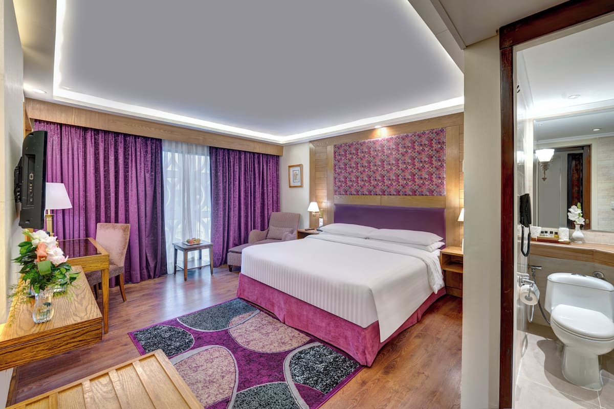 Une chambre d’hôtel moderne dotée d’un grand lit, d’un bureau avec une lampe, d’une chaise, d’une petite table et d’une salle de bain avec des toilettes et un lavabo.
