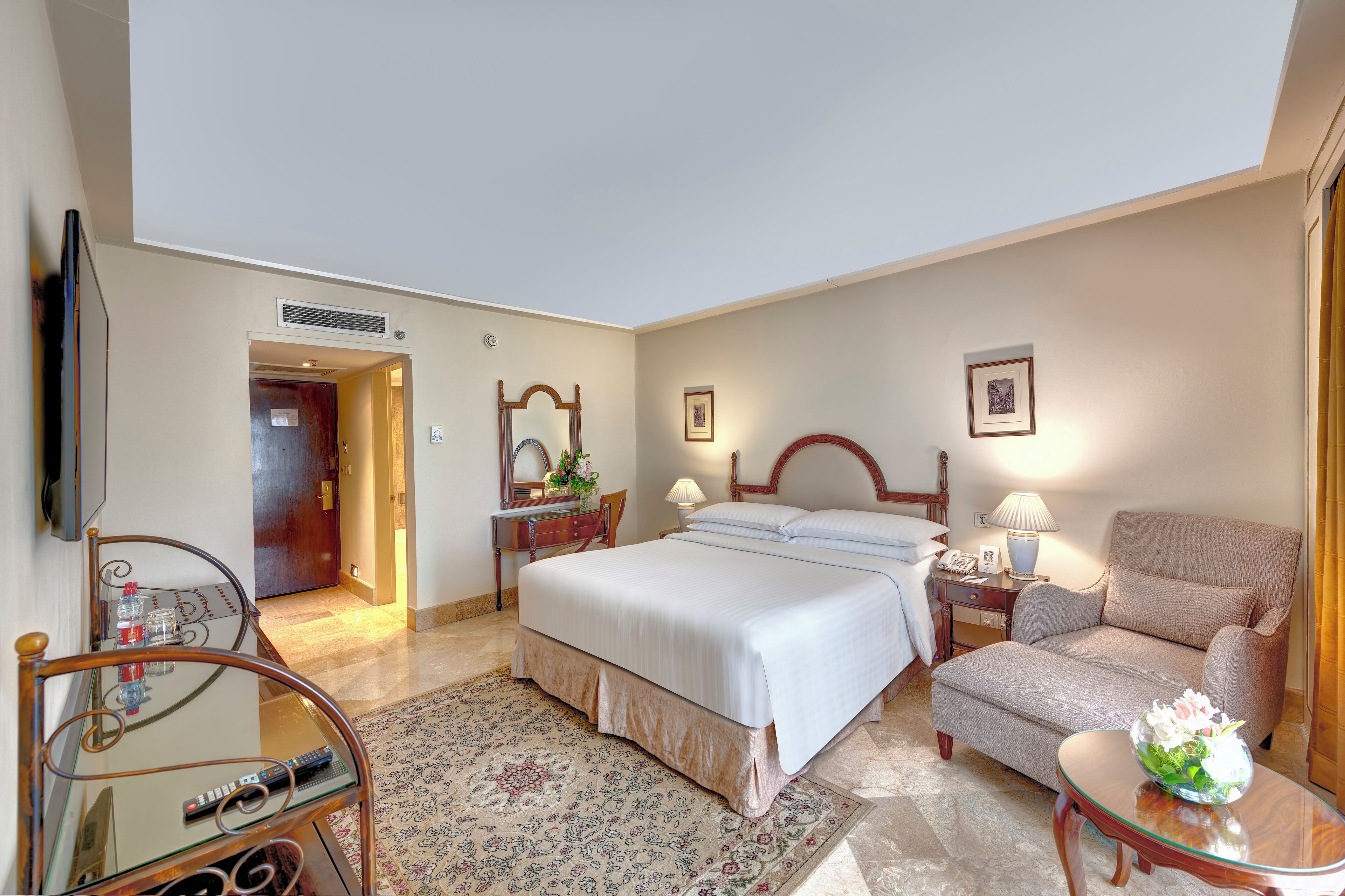 A hotel room with a bed, a chair, a desk with a lamp, a TV, and a decorative vase.