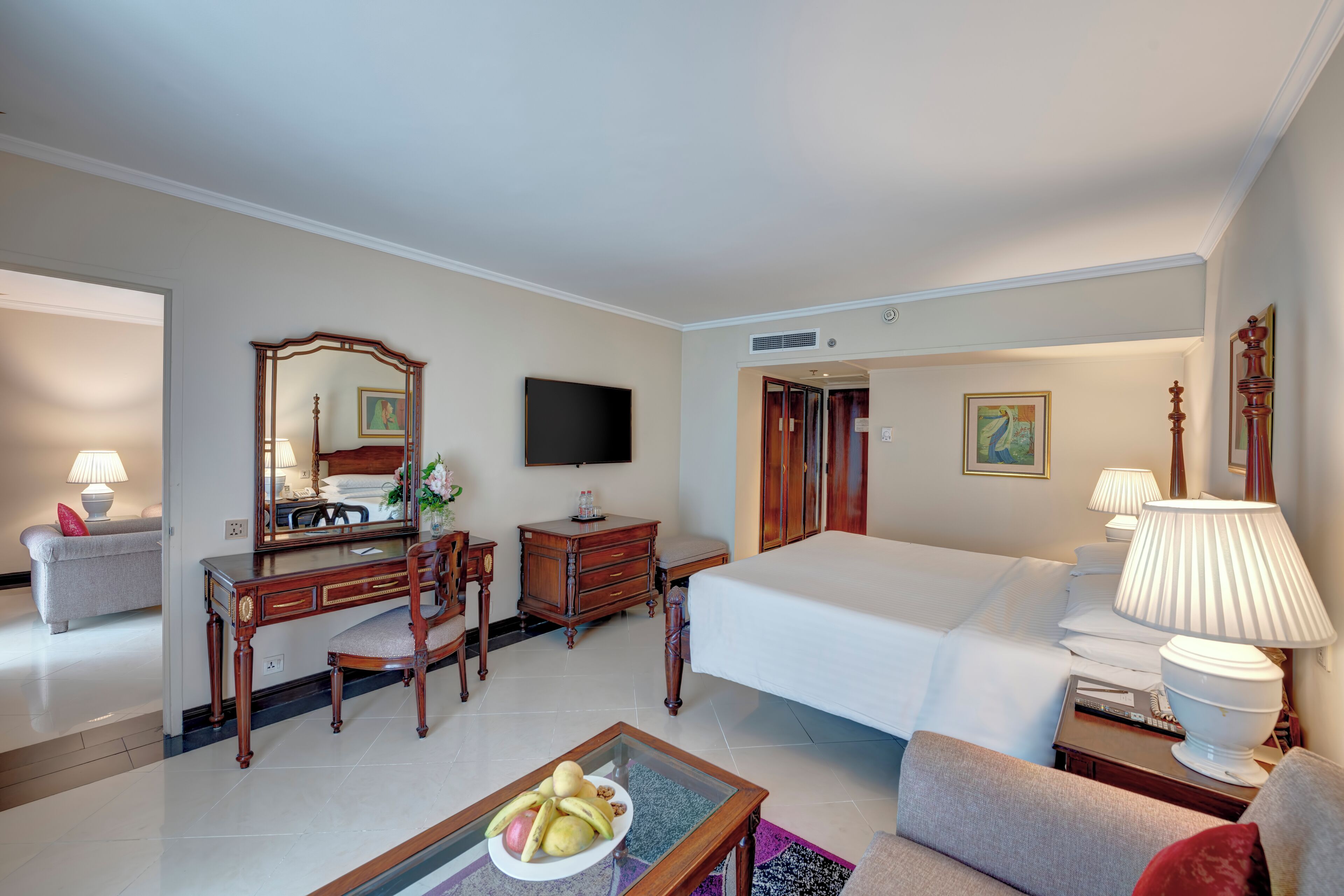 A hotel room with a bed, a desk with a chair, a television, a dresser, and a dining table with fruit.