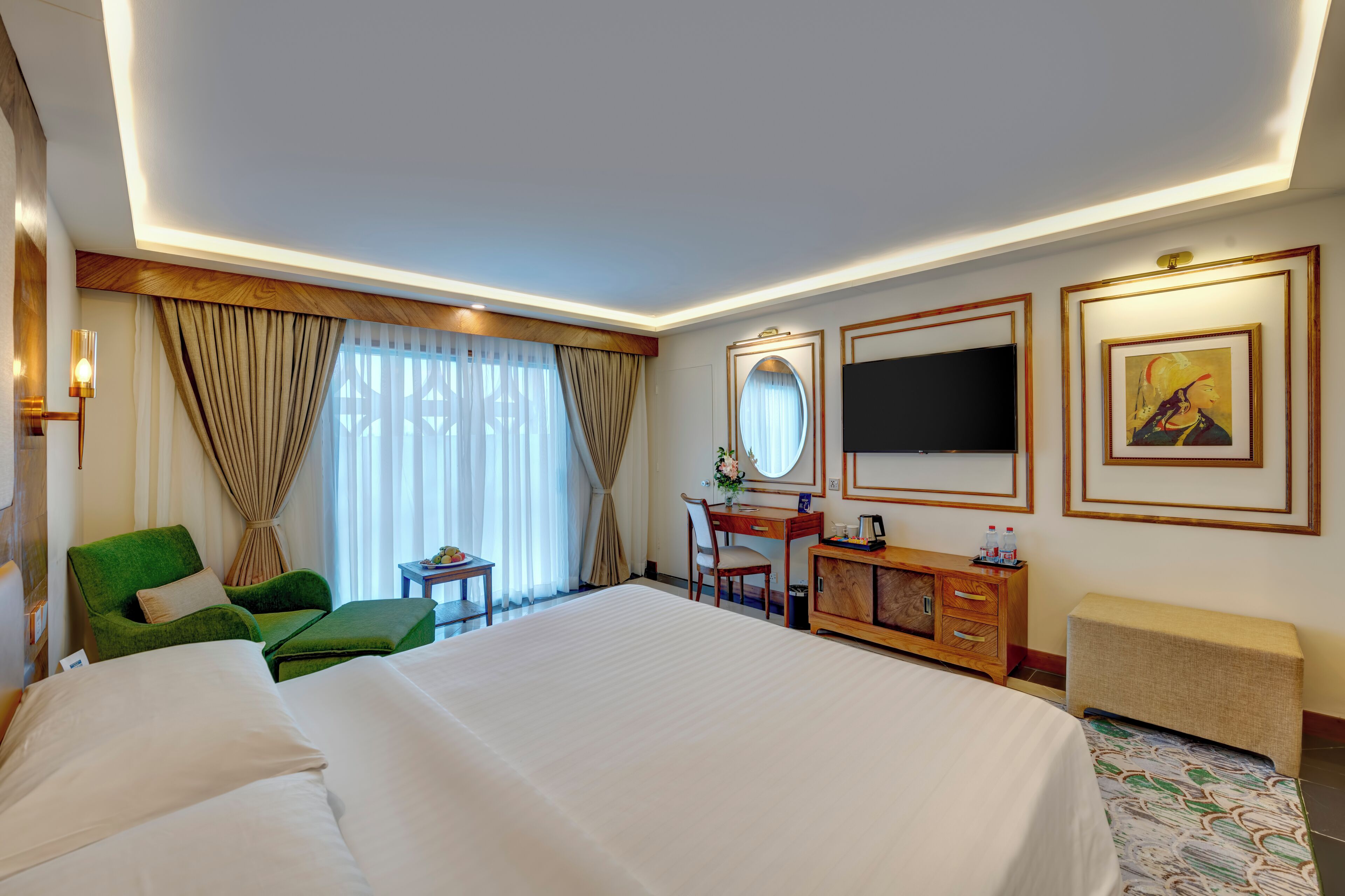 A hotel room with a large bed, a desk, a green armchair, a TV, and artwork on the walls.