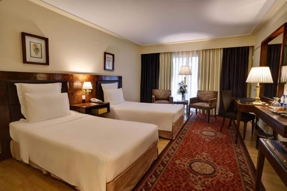 Une chambre d’hôtel avec deux lits, un bureau, une chaise, une lampe et une fenêtre avec des rideaux.