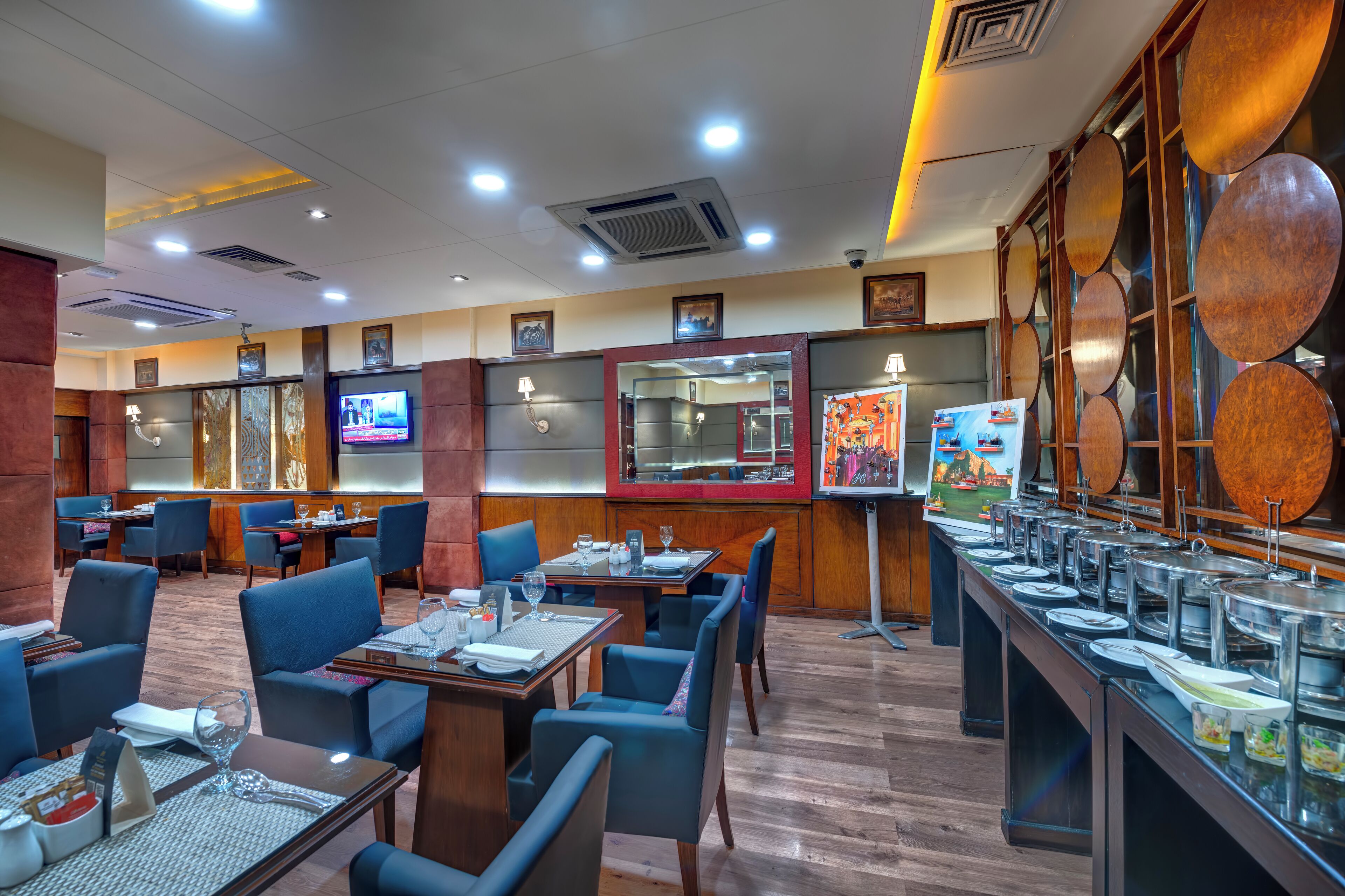 A restaurant with multiple dining tables and chairs, a buffet area with various food items, and a television screen on the wall.