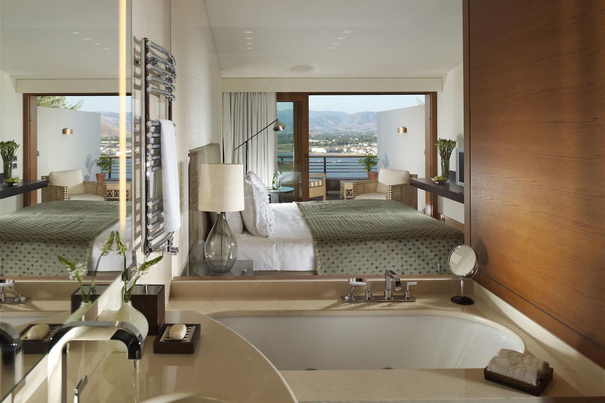 Ein modernes Hotelzimmer mit einem großen Bett, einer Badewanne, einem Schminktisch mit Spiegel und einem Balkon mit Meerblick.