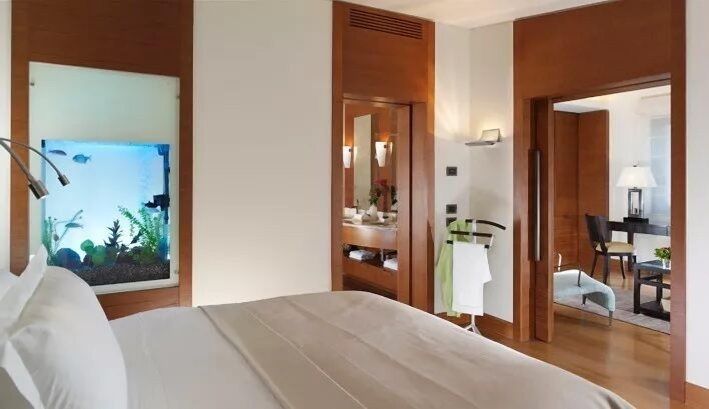 A hotel room with a bed, a large aquarium, a bathroom with a mirror, and a dining area visible through an open door.