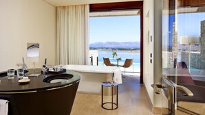 Een moderne hotelkamer met een keukenhoek, een eettafel met stoelen, een klein bijzettafeltje en een groot raam met uitzicht op een meer en bergen.