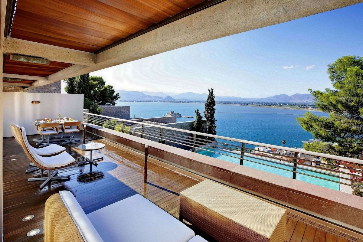 Ein Balkon mit Holzboden, weiß gepolsterten Sitzgelegenheiten und einem Tisch mit Stühlen. Im Hintergrund ist ein Pool zu sehen.