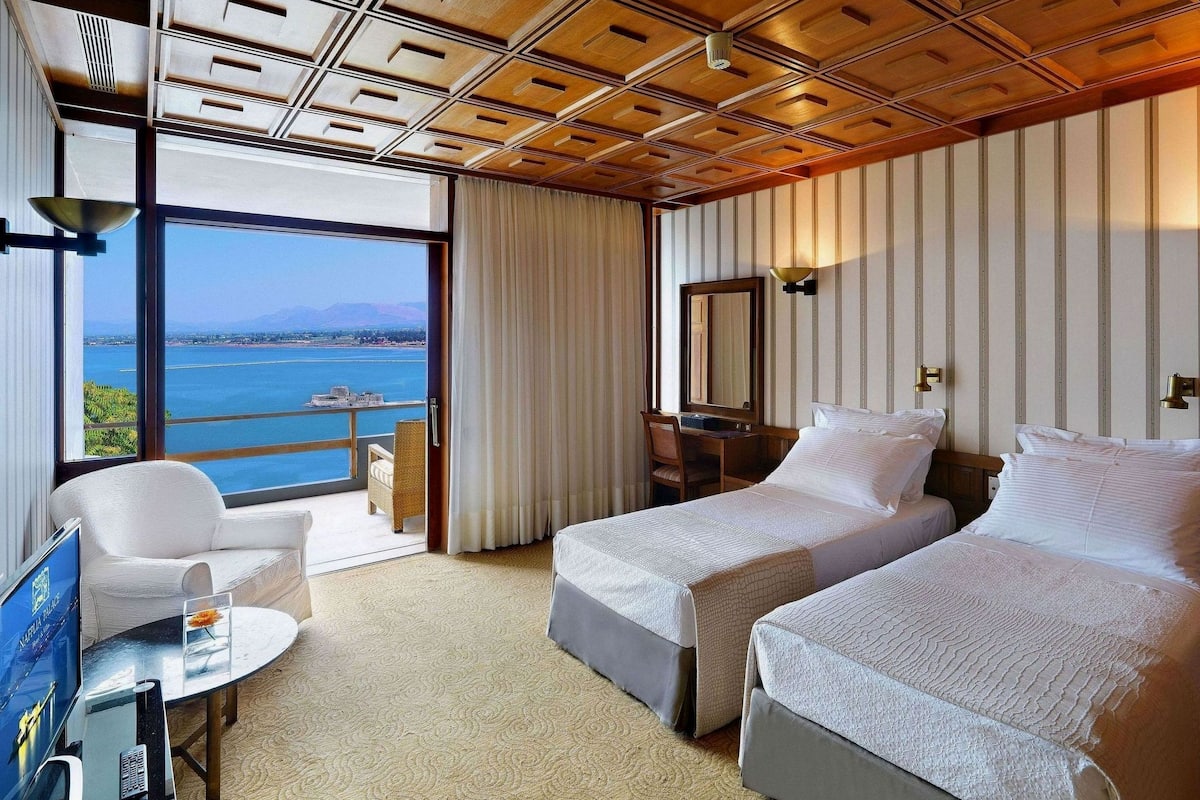 Ein Hotelzimmer mit zwei Betten, einem Schreibtisch, einem Sessel und einem großen Fenster mit Blick auf das Meer.