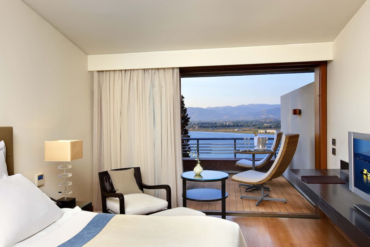 Ein Hotelzimmer mit Bett, zwei Sessel, einem kleinen Tisch, einem Fernseher und einem Balkon mit Blick auf Berge und Wasser.