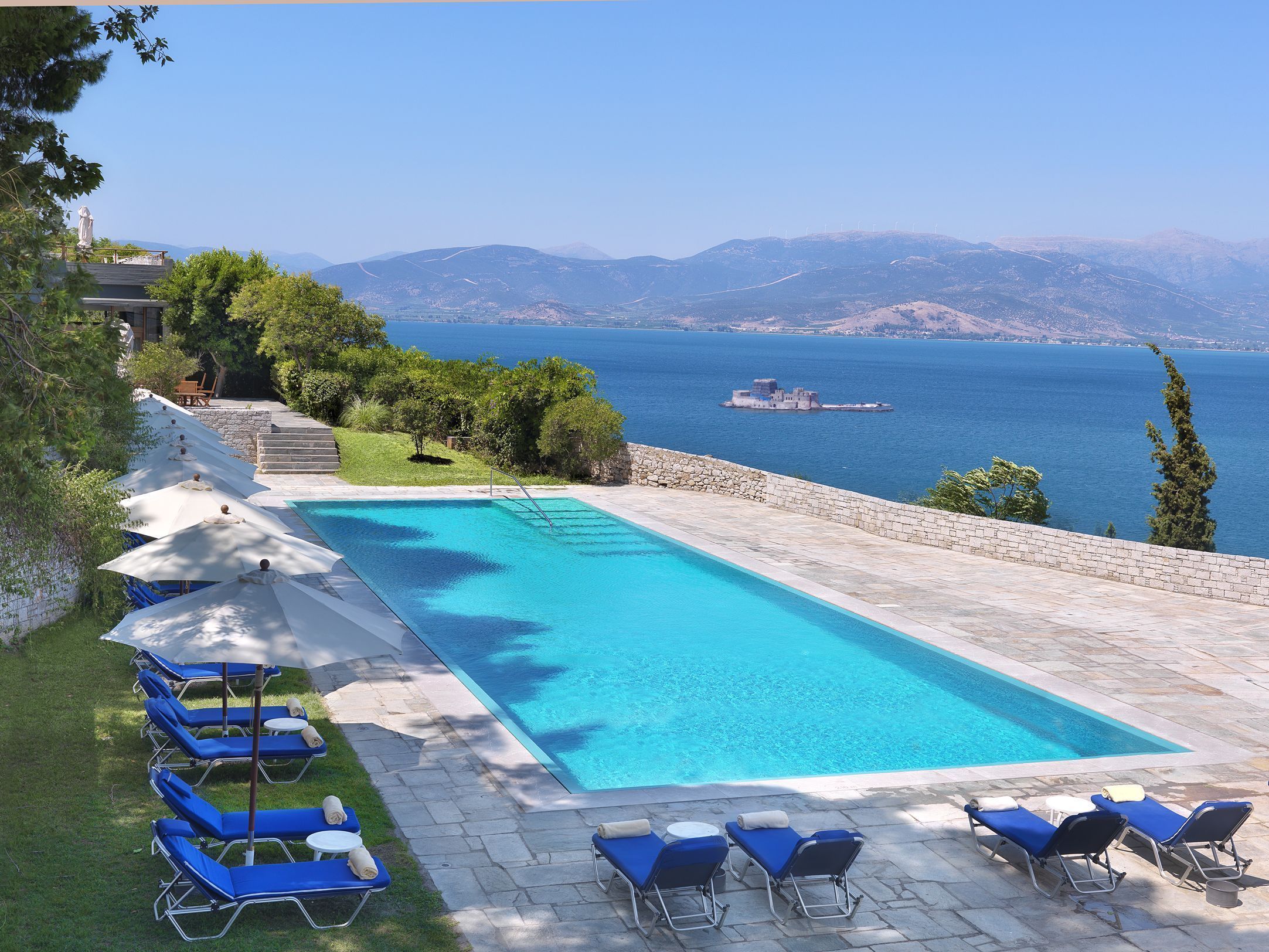 A swimming pool with lounge chairs and umbrellas, surrounded by a stone wall and greenery, overlooking a body of water and distant mountains.