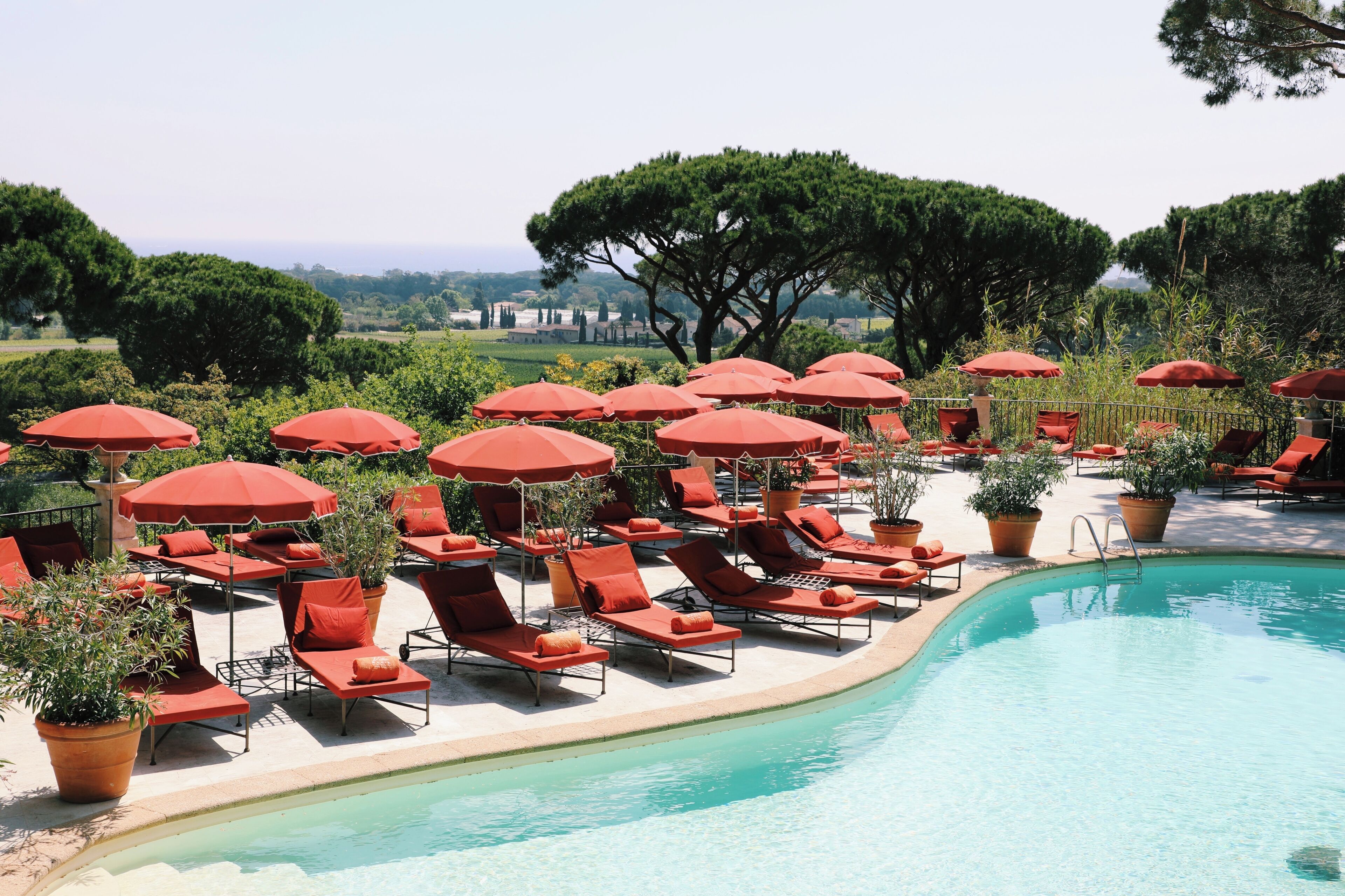 A poolside area with sun loungers and red umbrellas, surrounded by potted plants and trees.