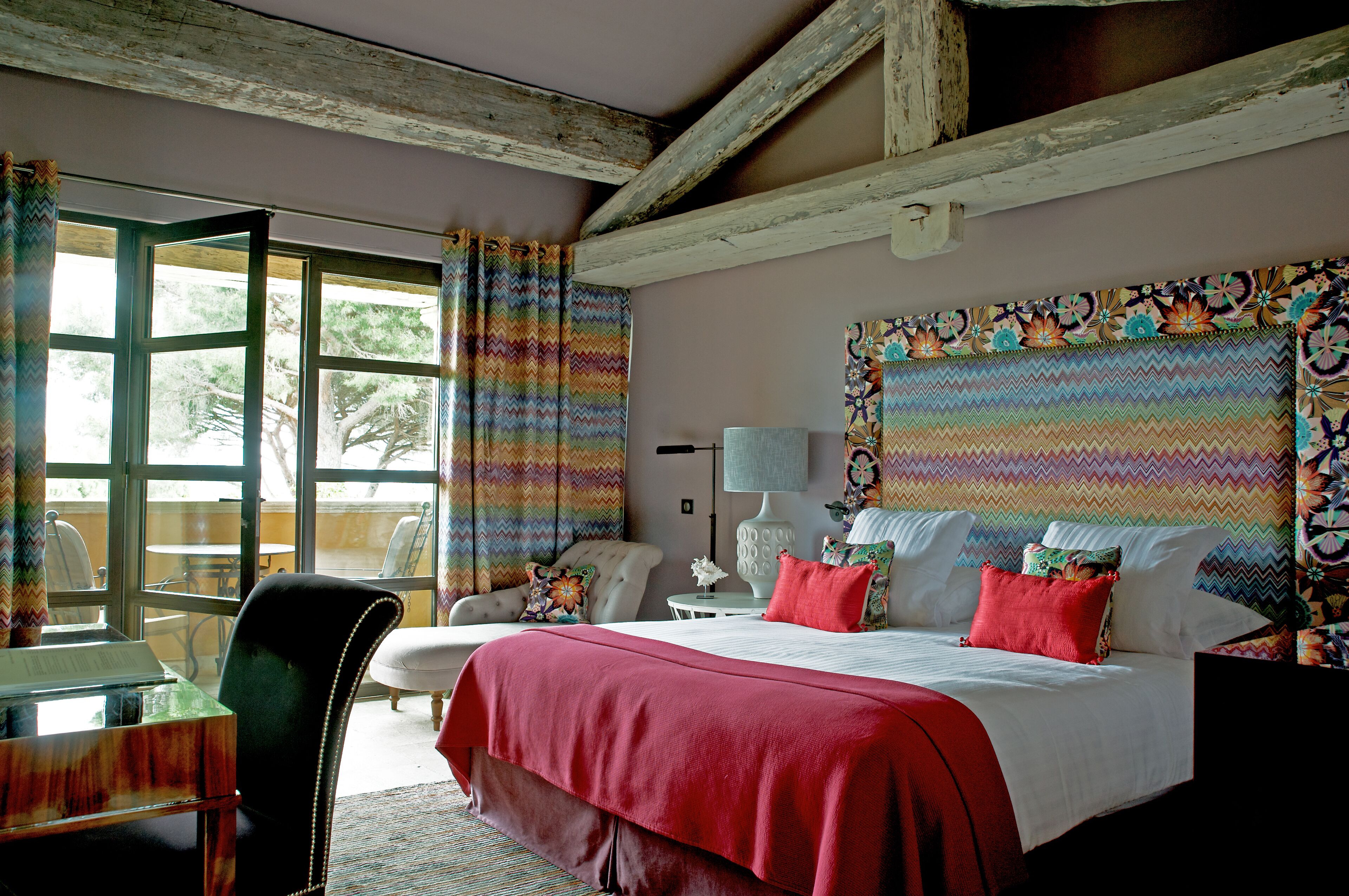 A bedroom with a large bed, a desk, and a chair. There is a window with curtains, a lamp, and a painting on the wall.