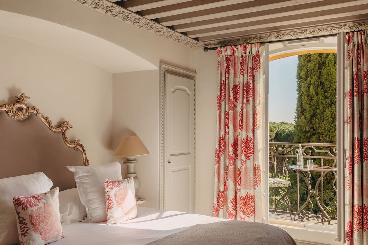 A bedroom with a bed, pillows, a lamp, a door, and a balcony with a table and chair.
