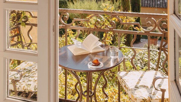 A balcony with a table set for two, a chair, and a view of a garden with trees and a hill in the background. - Hôtel Villa Marie St Tropez (Ramatuelle)