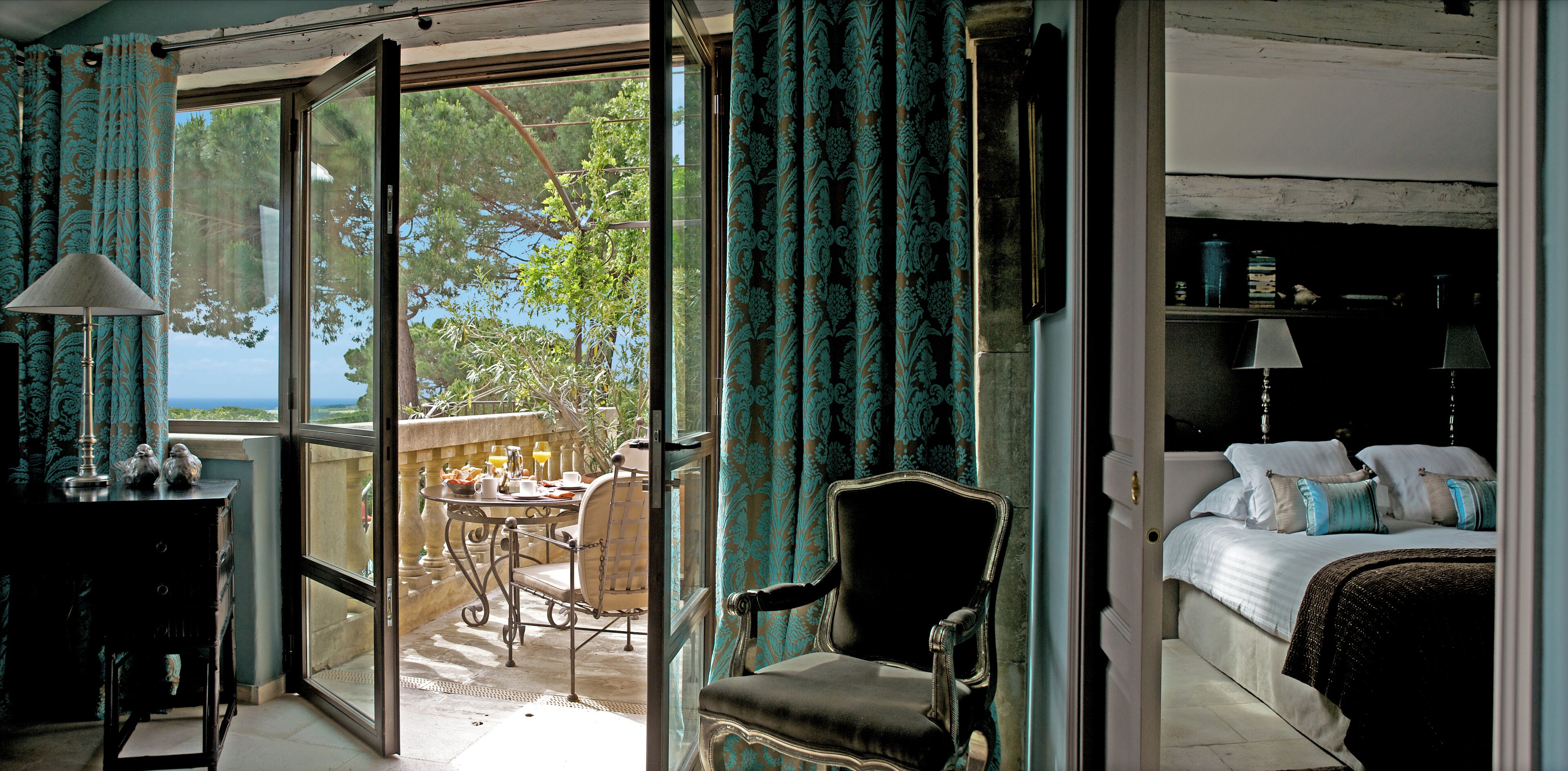 A room with a balcony, a bed, a chair, and a table with a lamp.