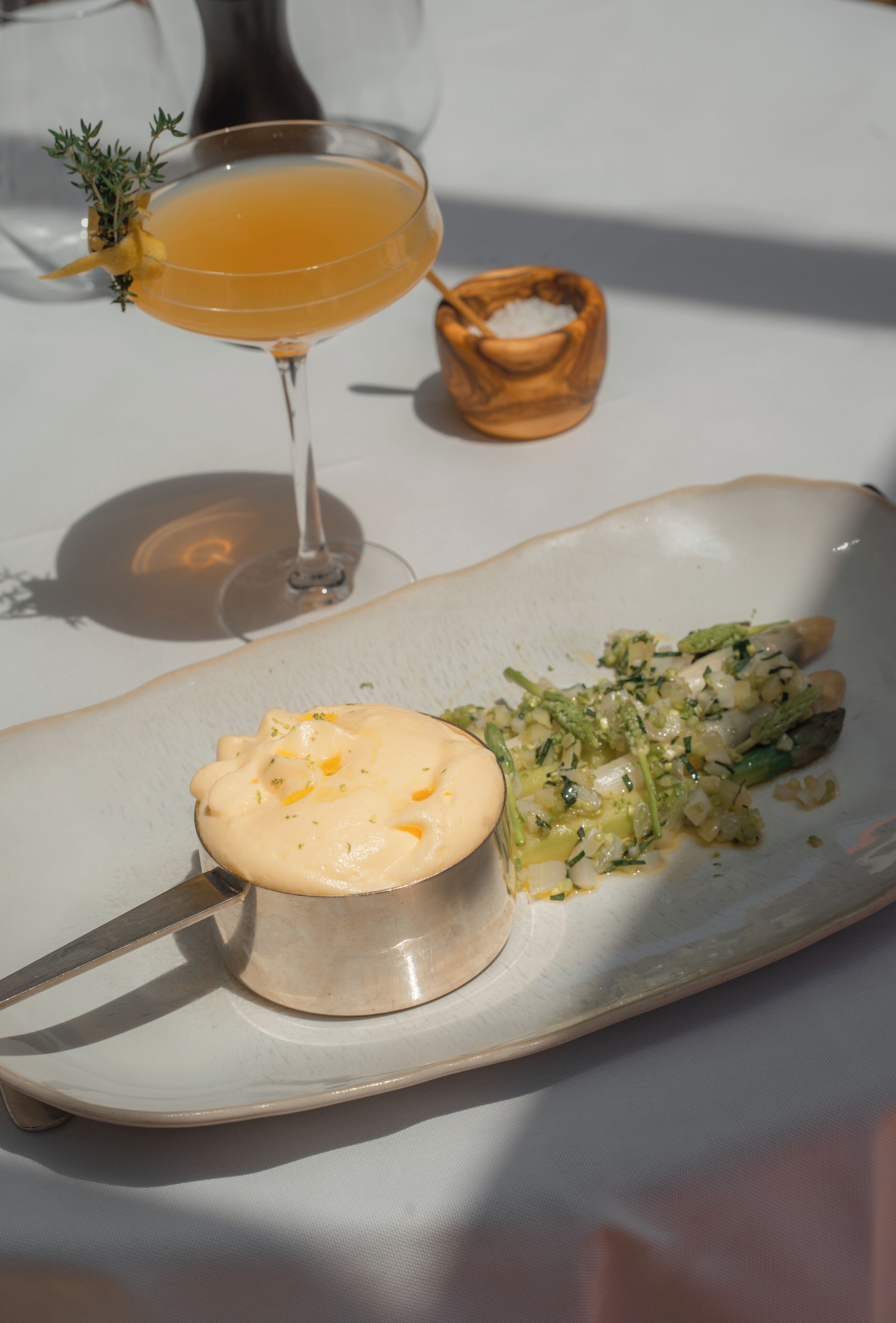 A plate with a creamy dish and a green vegetable side, a glass of orange liquid, and a small wooden bowl on a white table.