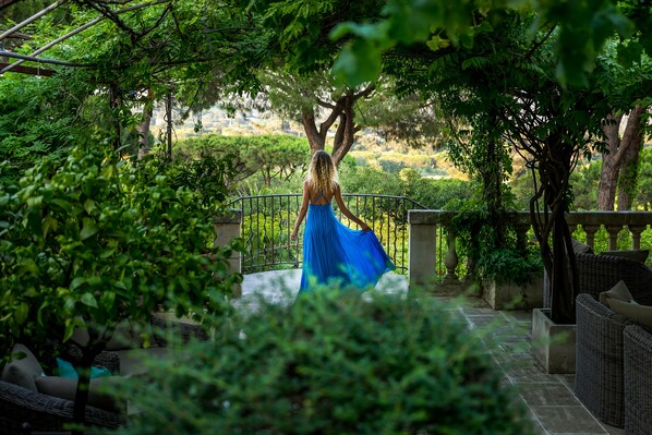 Une femme vêtue d’une robe bleue marche sur un sentier de pierres, entourée d’une végétation luxuriante et d’arbres.